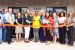 A group of people stand in front of a building with a sign and address 1404. A woman in the center cuts a large red ribbon with oversized scissors, while others smile and hold the ribbon, celebrating a grand opening.