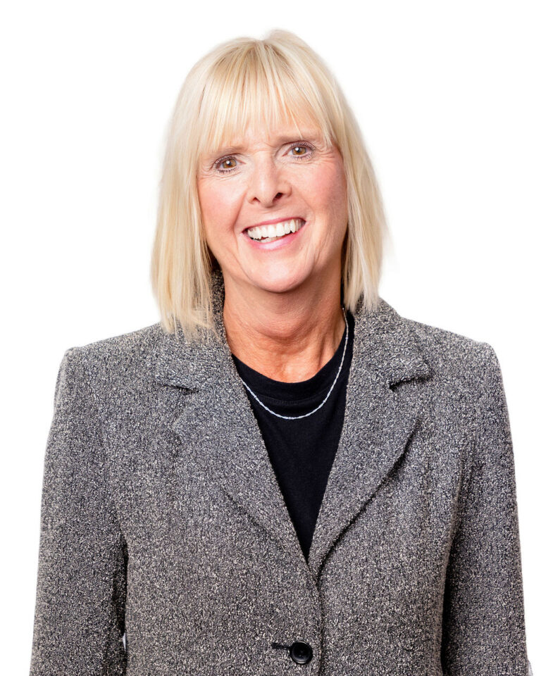 A smiling woman with straight blonde hair wears a gray blazer over a black top, standing against a white background.